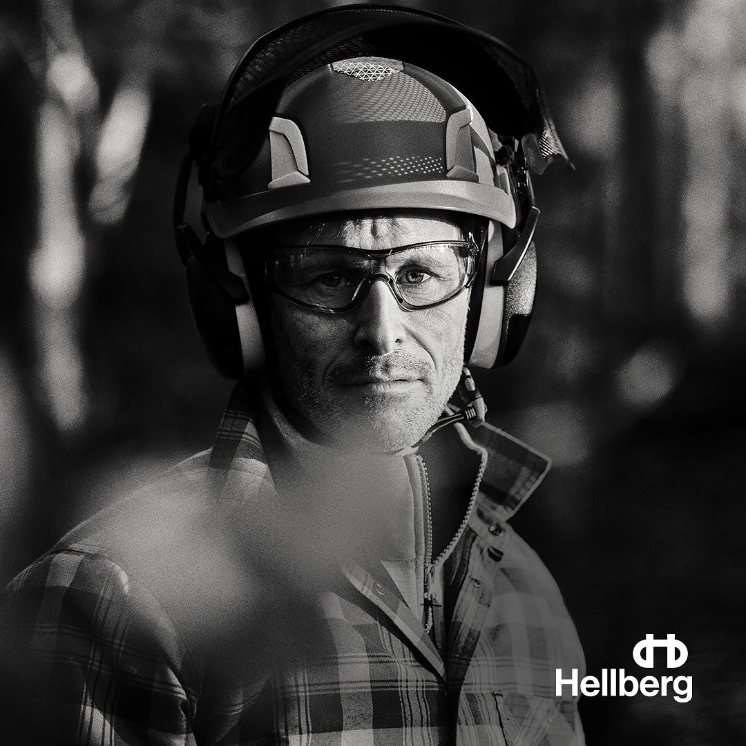 Mand med høreværn, sikkerhedshjelm og briller i skoven med Hellberg logo